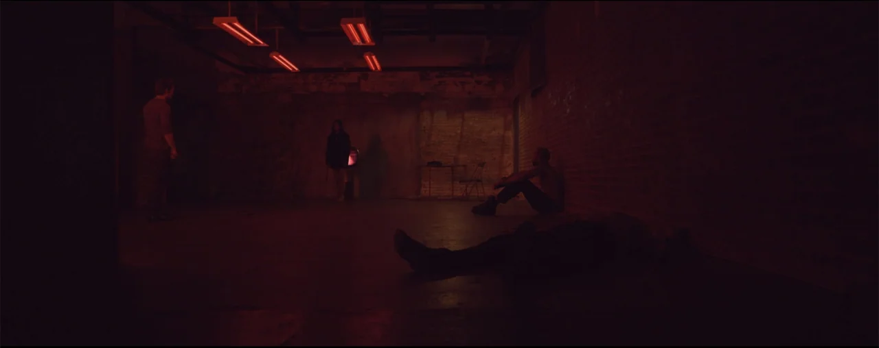 El Último Juego (2025) movie still: wide — An industrial room features exposed brick walls and red overhead fluorescent lights. Three people are…