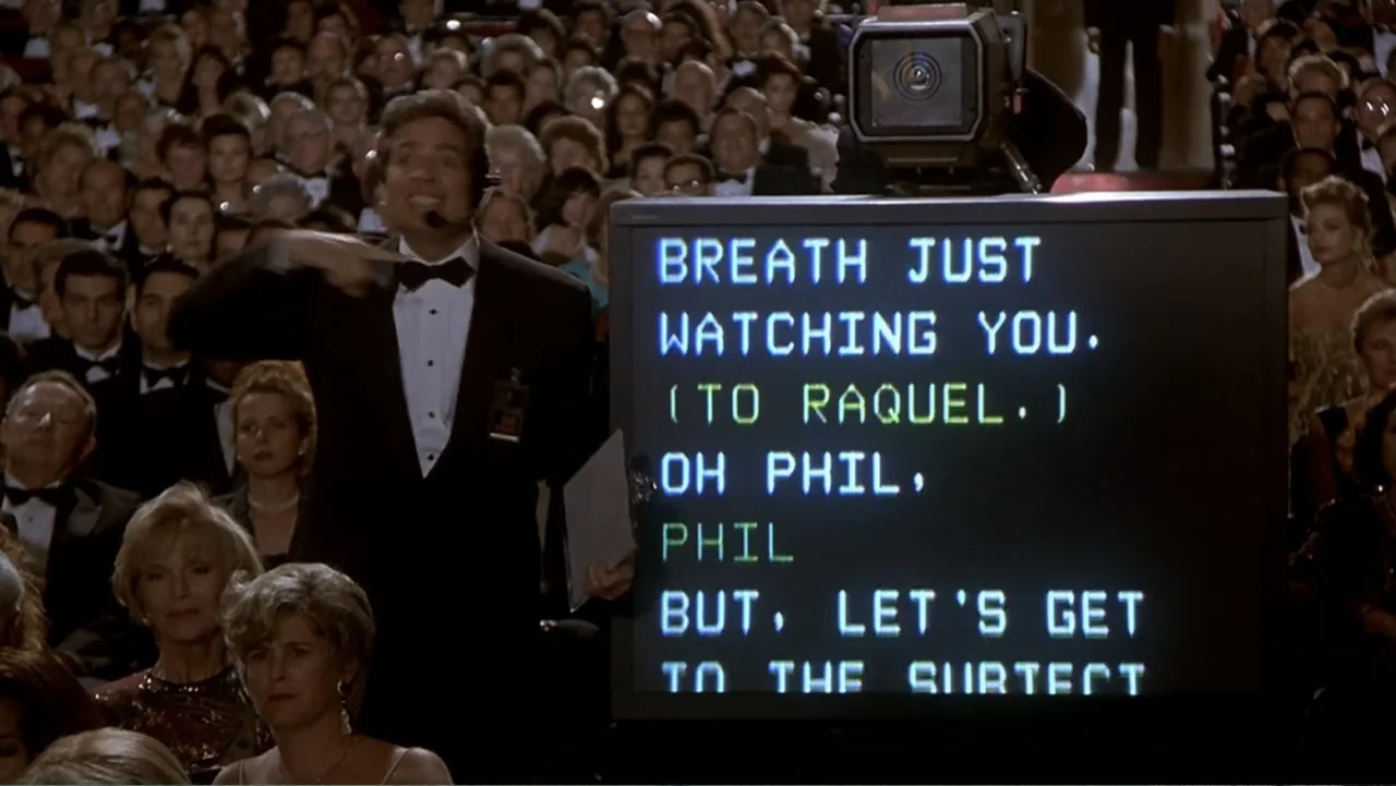 Naked Gun 33⅓: The Final Insult (1994) movie still: medium — A man in a tuxedo stands beside a large teleprompter in a dark theater auditorium. He…