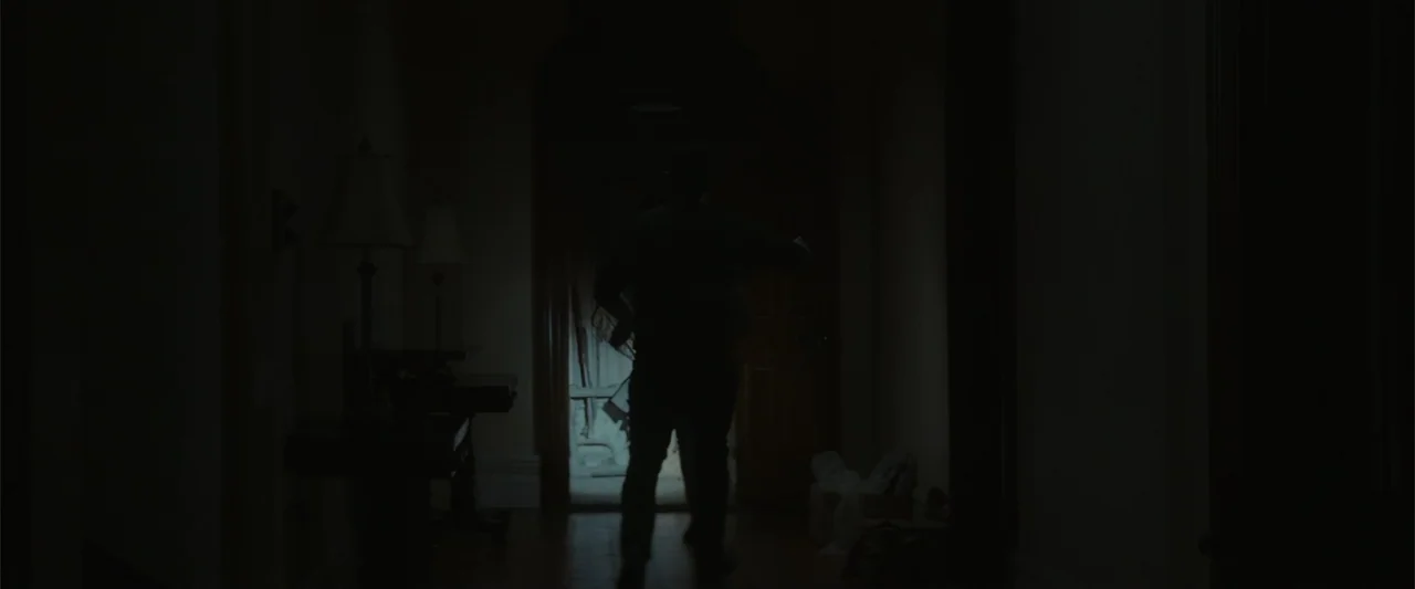 The Cellar (2022) movie still: fullbody — A dark residential hallway shows silhouettes of furniture and a lit doorway in back. A man walks through…