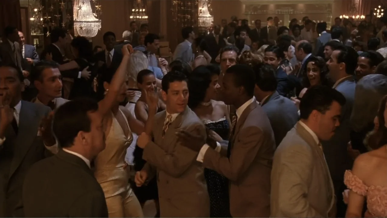 The Mambo Kings (1992) movie still: medium — A crowded formal ballroom interior filled with many people in 1950s evening attire. A group of men and…