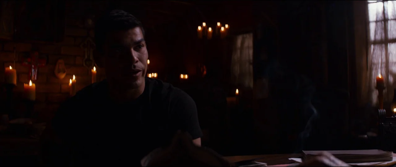 El Chicano (2018) movie still: medium — A dark room illuminated by several glowing candles set against a dark background. A man sits at a table in…