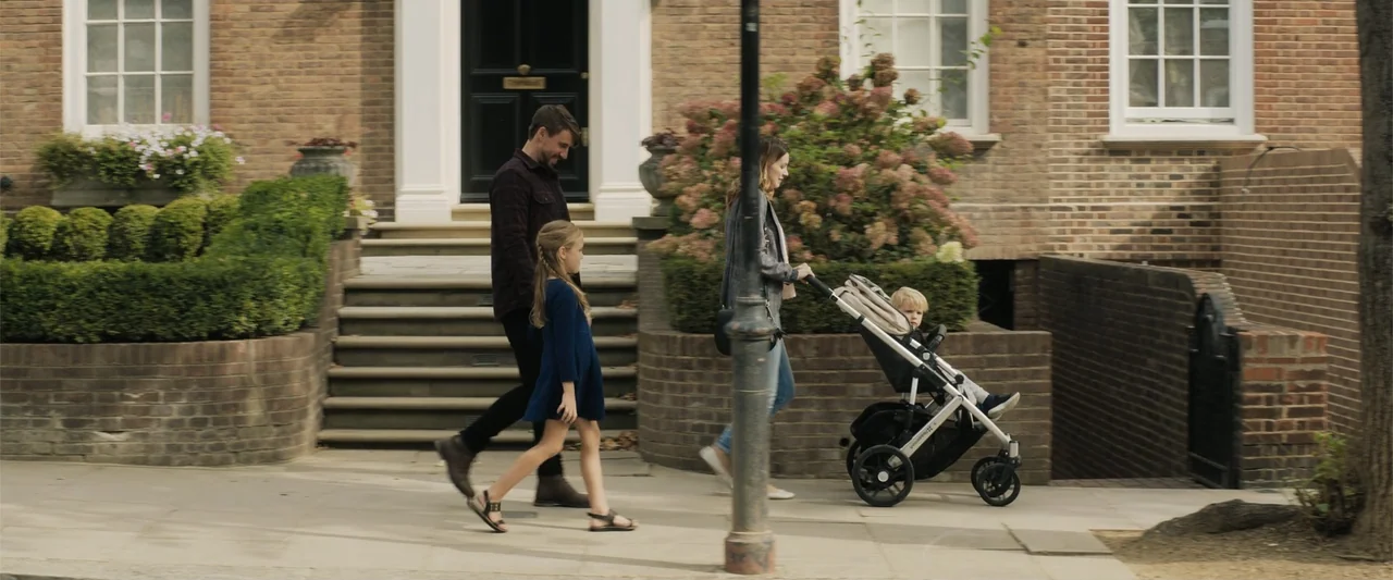 The 355 (2022) movie still: fullbody — A suburban residential street features red brick townhouses with steps leading to black doors. A family of…