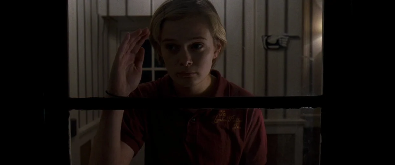 The Innkeepers (2011) movie still: medium — The dimly lit interior shows a wood-paneled lobby with a vintage directional sign on the wall. A young…
