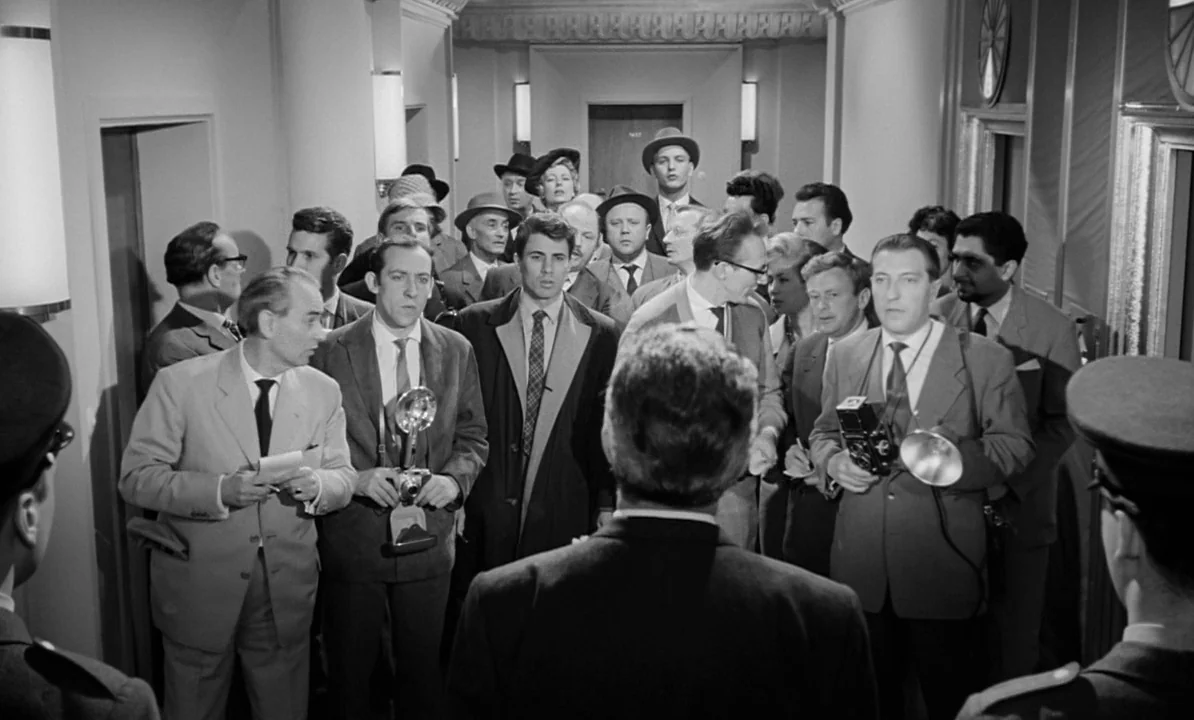 The 1,000 Eyes of Dr. Mabuse (1960) movie still: medium — A crowded, narrow hotel corridor lined with sconces is filled with men in suits. The group…