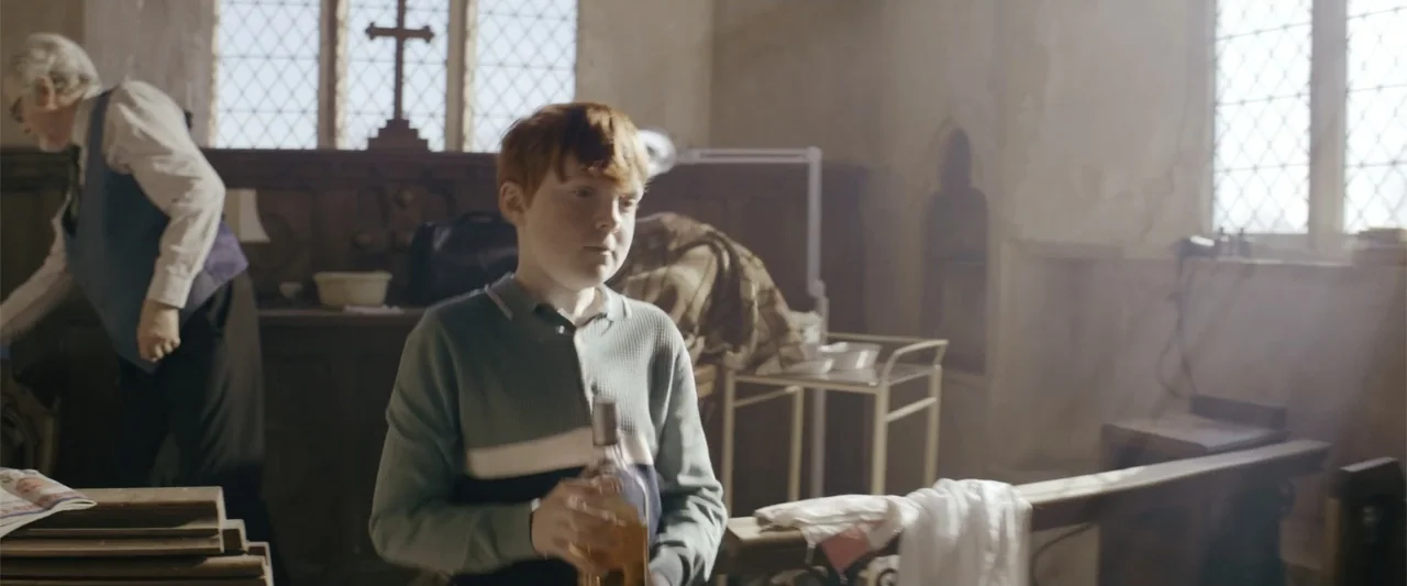 One Ranger (2023) movie still: medium — A church interior with wooden pews, stone walls, and leaded glass windows. A red-haired boy holds a glass…