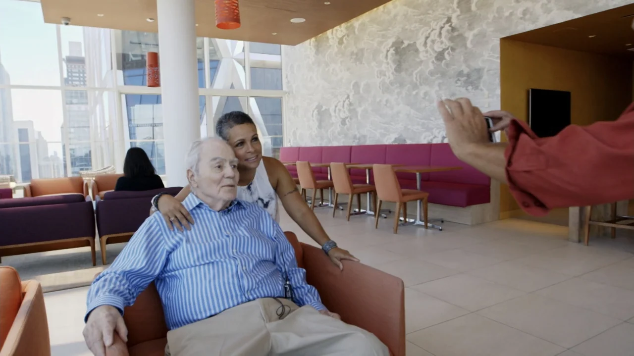 Breslin and Hamill: Deadline Artists (2018) movie still: medium — A sunlit modern lobby features patterned wallpaper and rows of purple armchairs. A…