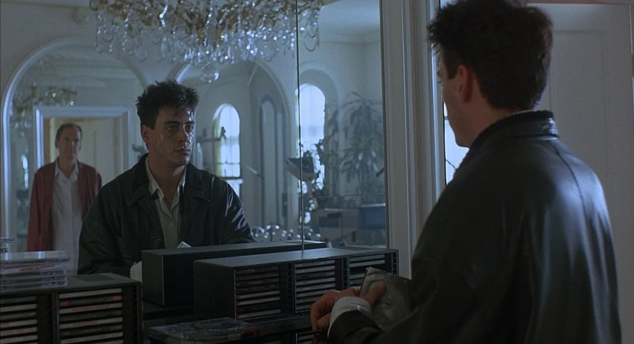 Less Than Zero (1987) movie still: medium — A room with large mirrored walls and an ornate crystal chandelier overhead. A young man stands in front…
