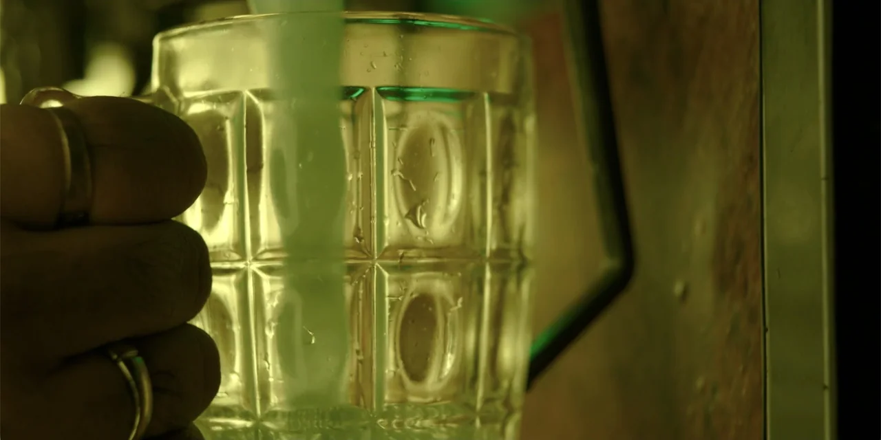 Maja Stoneface (1996) movie still: closeup — A dimly lit bar setting with a textured glass mug in the foreground. A person's hand with a ring holds…