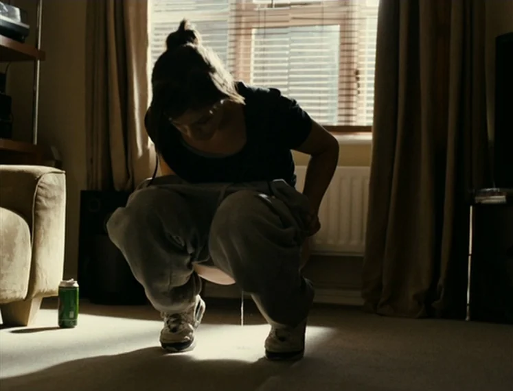 Fish Tank (2009) movie still: medium — A dimly lit room with carpeted floors and closed curtains over a window. A woman in sweatpants crouches low…
