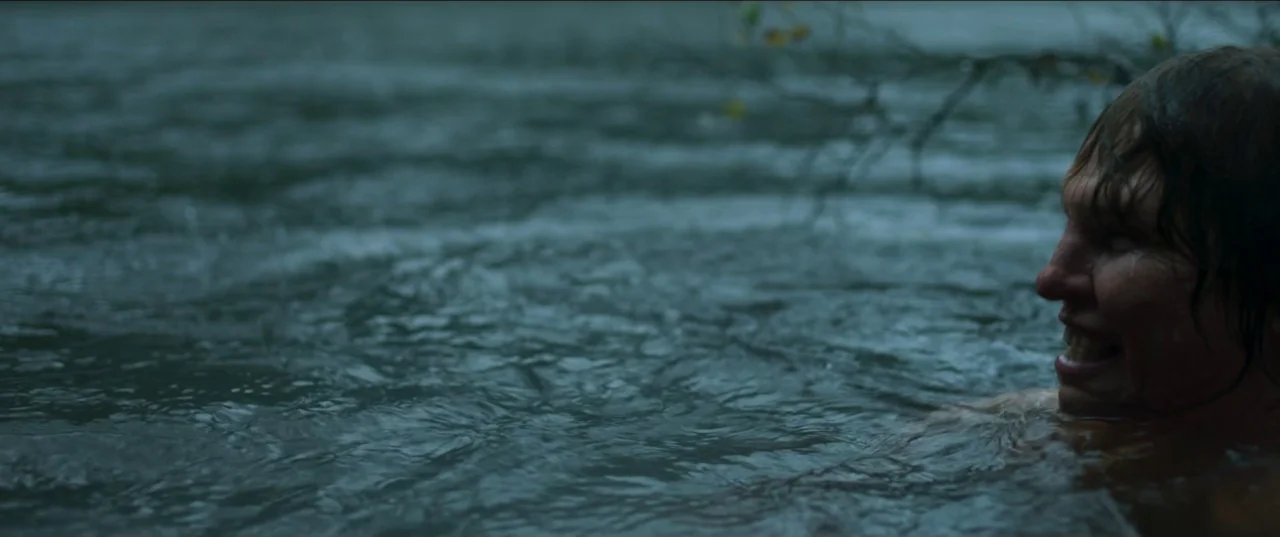 Border movie still: closeup — A body of murky, dark water fills the frame with a branch slightly visible. A man is partially submerged in the water…