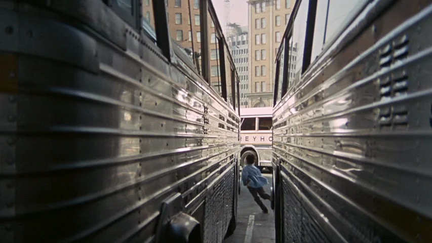 Petulia (1968) movie still: wide — A narrow gap between two silver Greyhound buses in a crowded terminal. A young boy runs quickly through the…