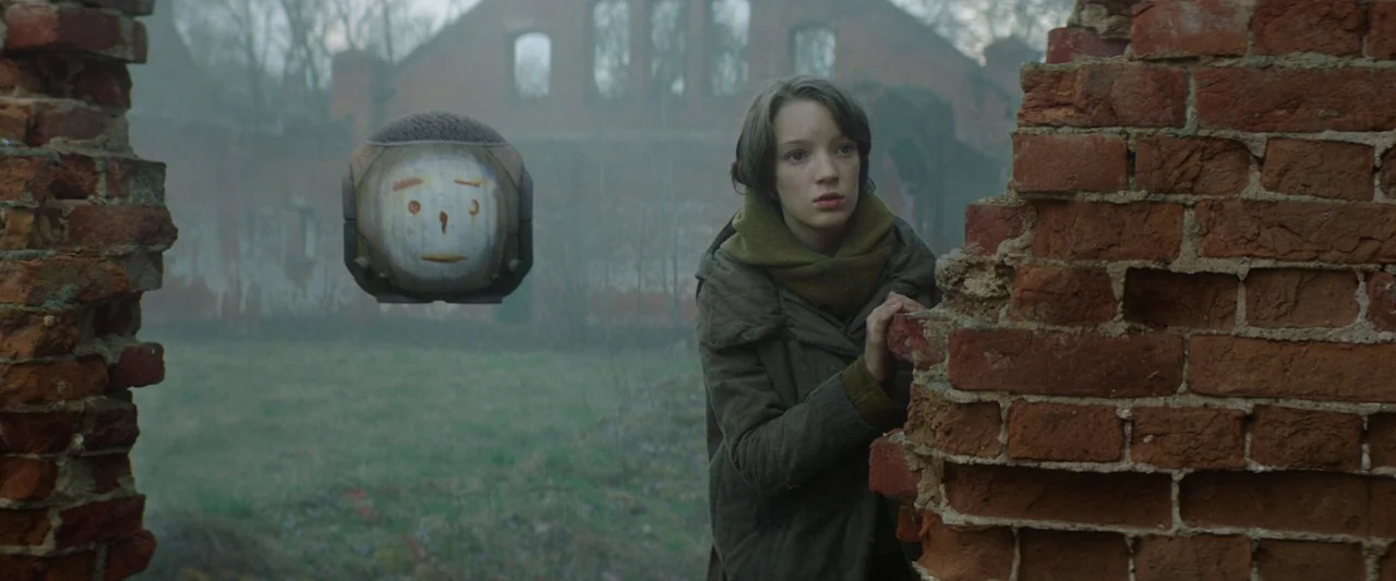 Vesper (2022) movie still: medium — An open expanse features a ruined brick structure under a thick grey mist. A woman hides behind a fragmented…