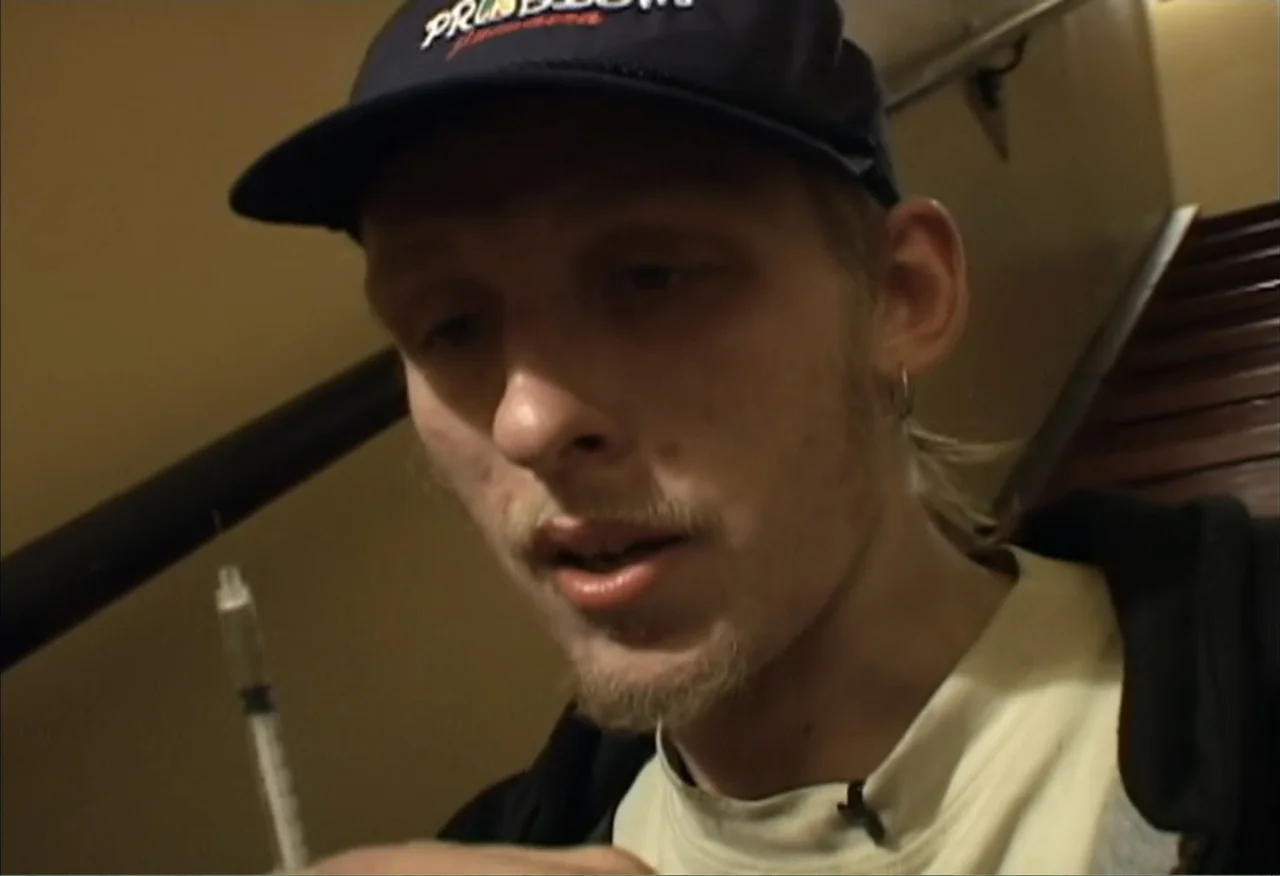 Dope Sick Love (2005) movie still: closeup — A dimly lit indoor stairwell features a dark railing and muted yellow wall paint. A young man holds a…