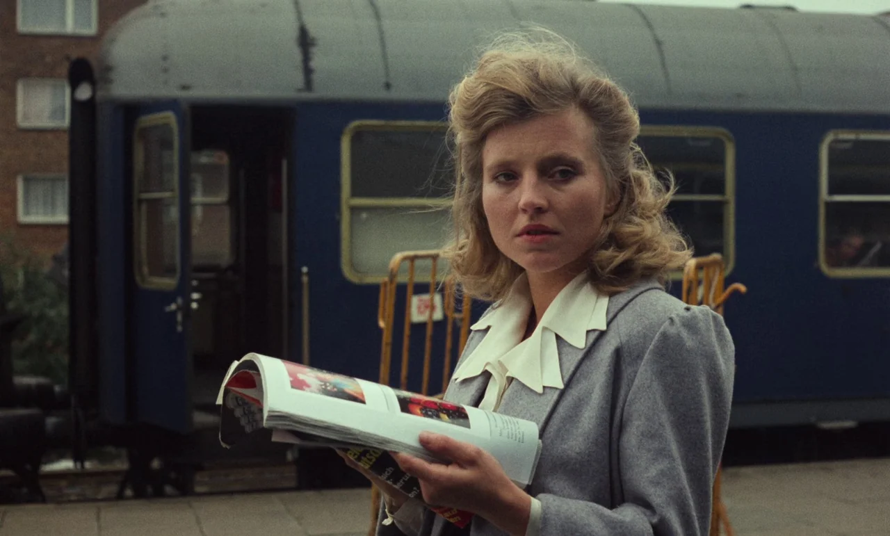 Wrong Move (1975) movie still: medium — A railway platform features a parked blue passenger train car in the background. A woman in a grey blazer…
