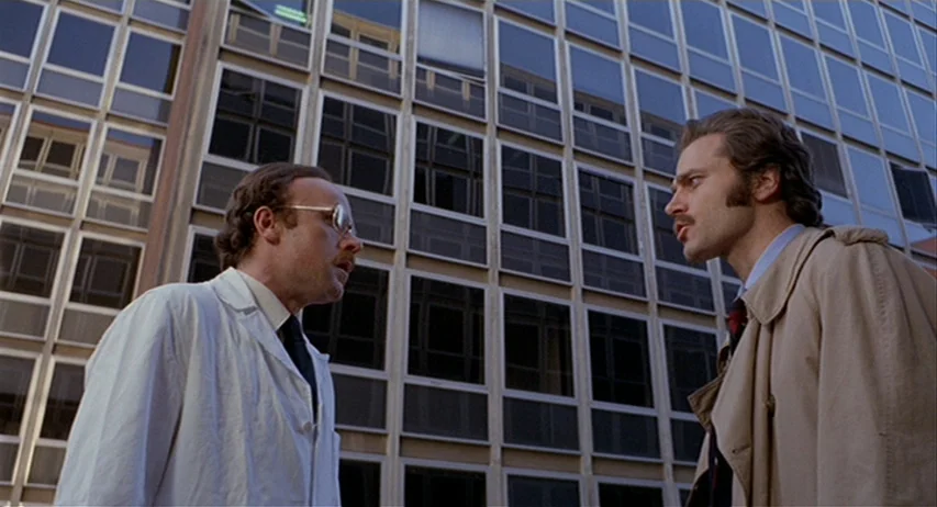 The Fifth Cord (1971) movie still: medium — Two men stand in front of a modern glass-paneled building with steel frames. The men face each other…