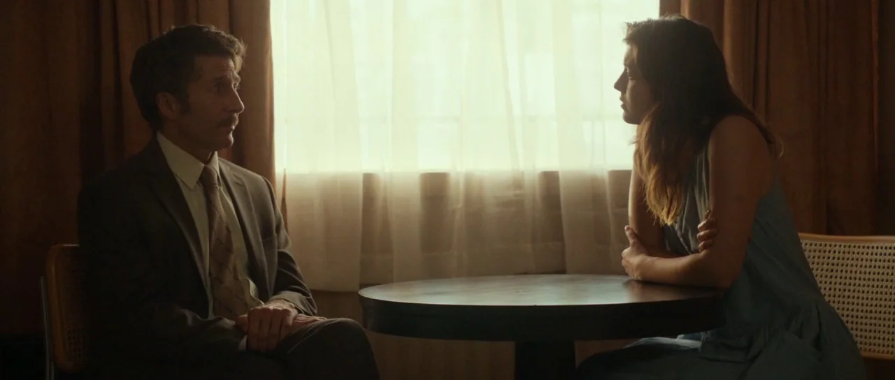 Faults (2014) movie still: medium — Two individuals sit at a small circular table in front of a draped window. The room features dark wood furniture…