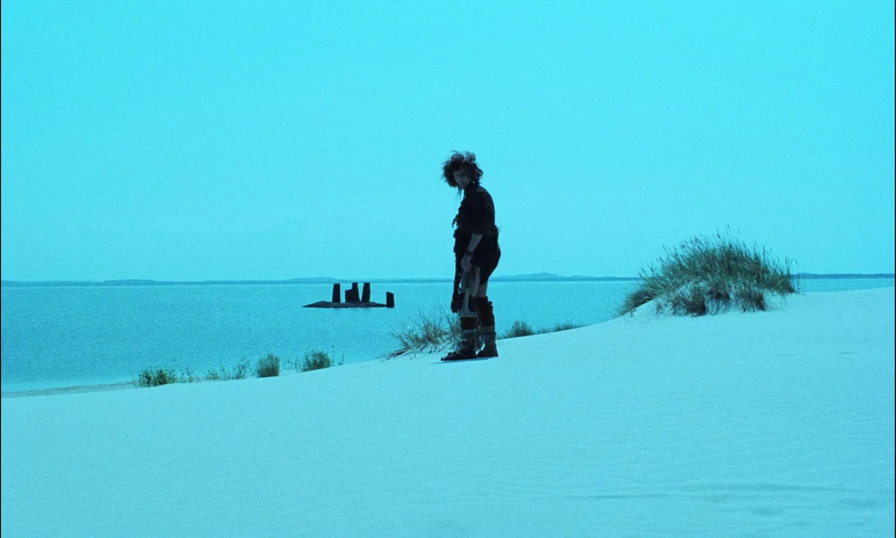 On The Silver Globe (1988) movie still: wide — A figure stands upon a bright white sand dune overlooking a calm blue body of water. A submarine…