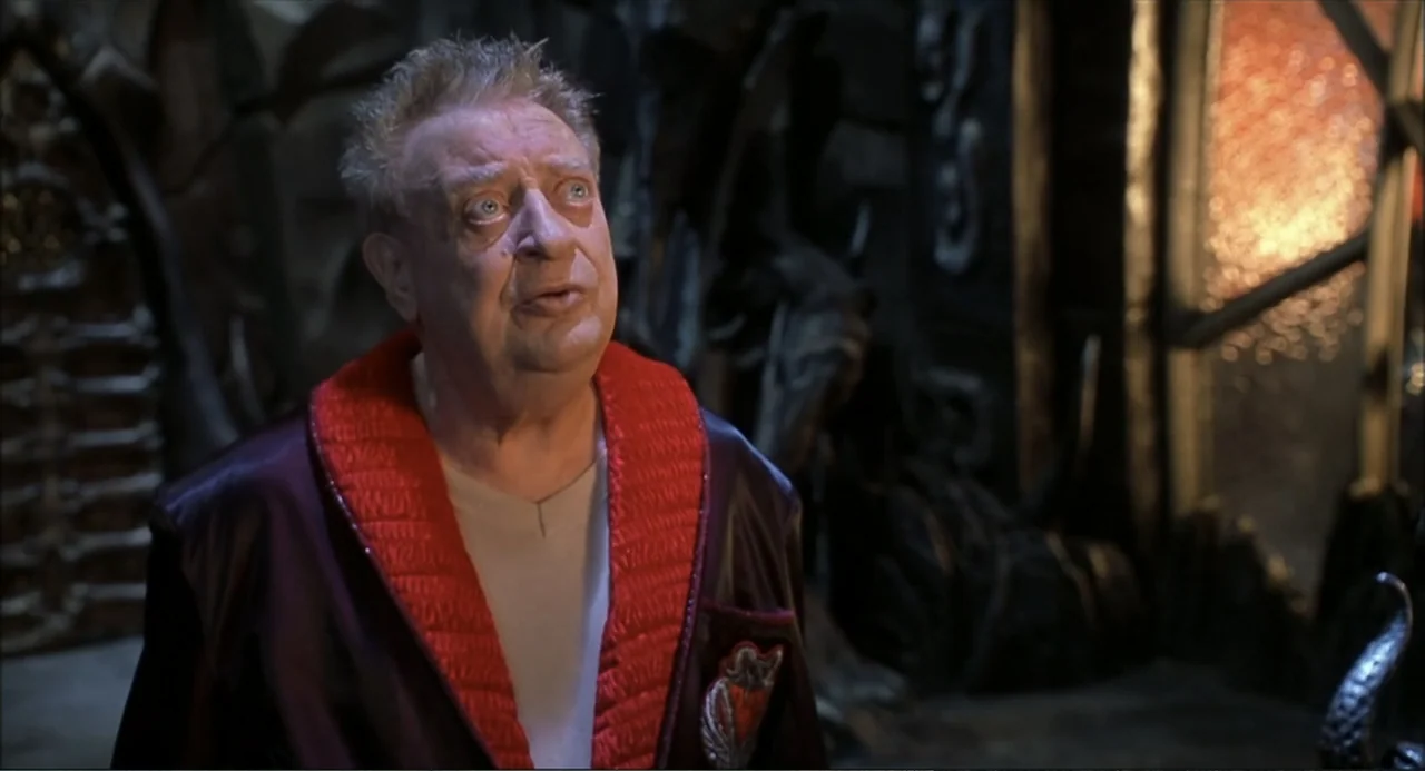 Little Nicky (2000) movie still: medium — An interior stone chamber features complex carved walls and dim ambient lighting. An elderly man in a dark…