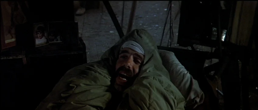 MASH (1970) movie still: closeup — A man lies within a makeshift shelter constructed of dark material and wooden supports. He is wrapped in an olive…