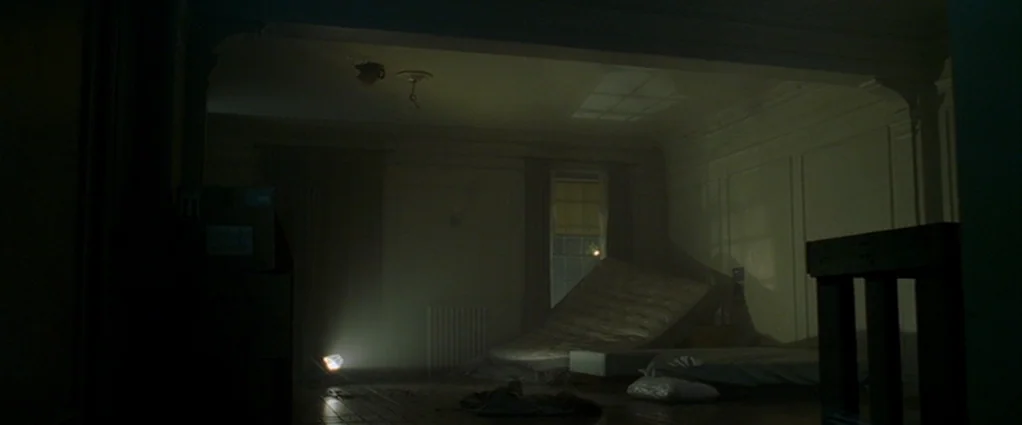Panic Room (2002) movie still: wide — A dark, sparsely furnished room features two mattresses on the floor and hazy air. A portable work light on…