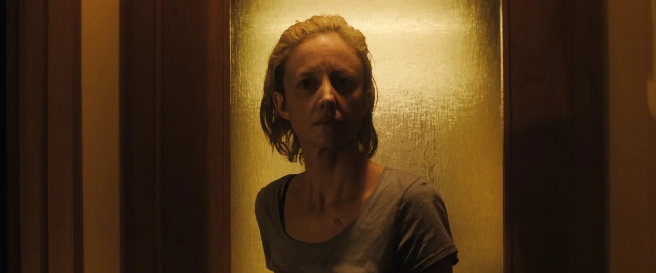 The Grudge (2020) movie still: medium — A woman stands in a dim doorway against a bright, illuminated yellow panel. She looks forward while light…