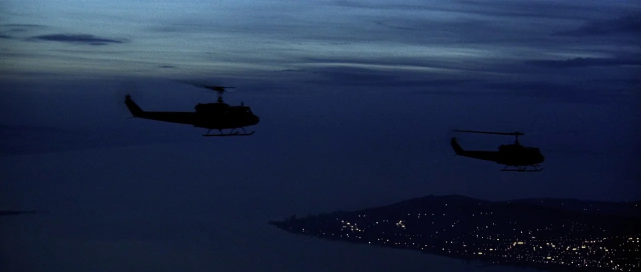Spy Game (2001) movie still: wide — Two helicopters fly in silhouette against a dark, twilight sky over a coastline. A dark landmass with scattered…