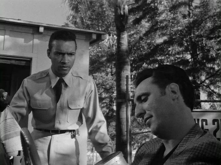 The Killing (1956) movie still: medium — An outdoor location featuring a light-colored building and a chain-link fence with foliage. A soldier in…
