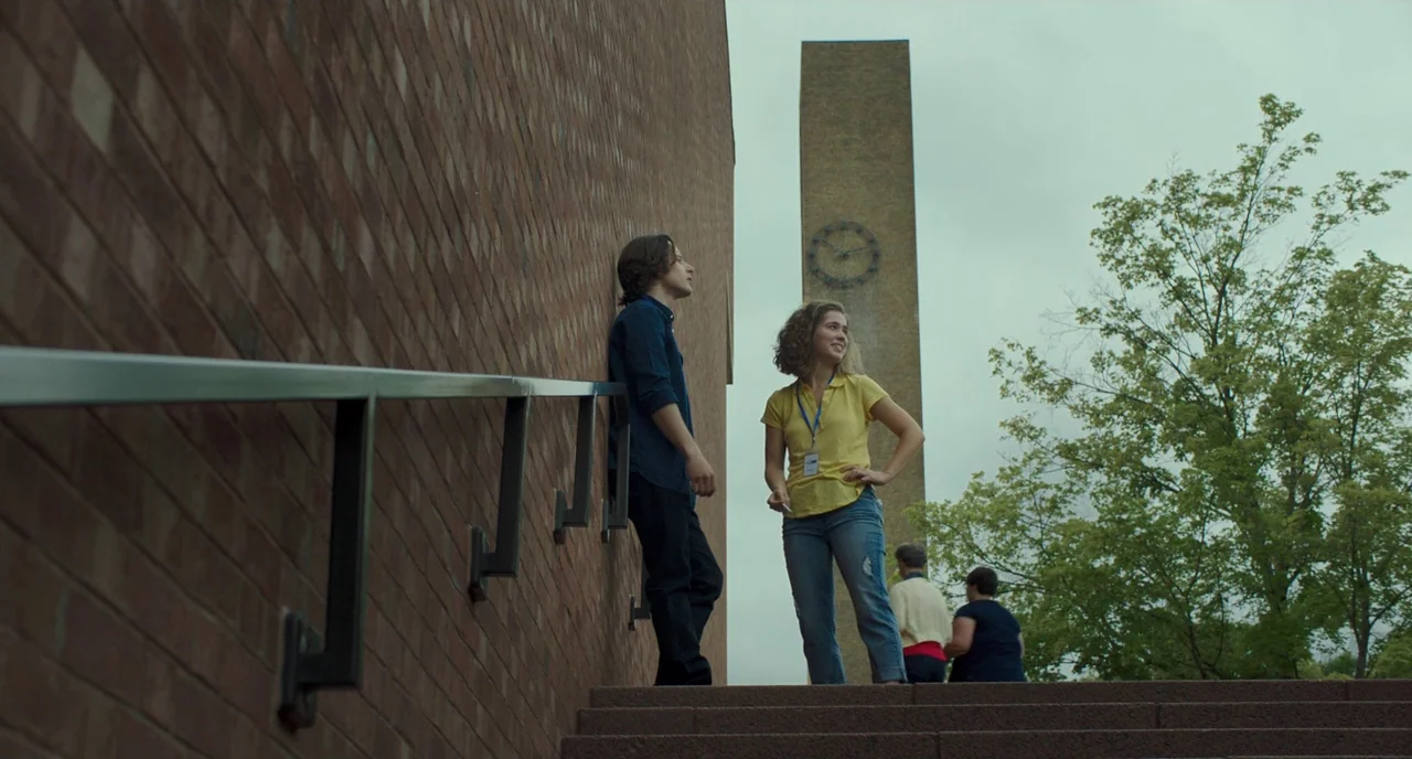 Columbus (2017) movie still: wide — Two individuals stand at the top of concrete stairs beside a tall brick structure. A modern clock tower rises in…