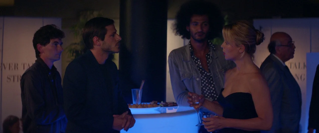 Sibyl (2019) movie still: medium — A dimly lit room contains a circular bar illuminated by a bright blue light. Several adults stand around the bar…
