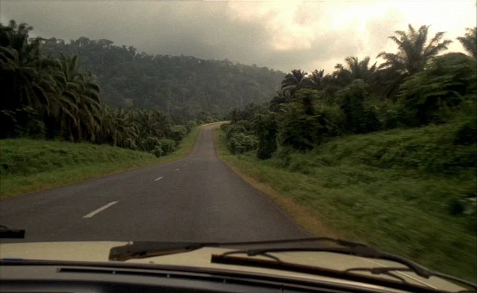 Chocolat (1988) movie still: establishing — A paved road cuts through a dense tropical landscape with palm trees and hills. The view is from inside…