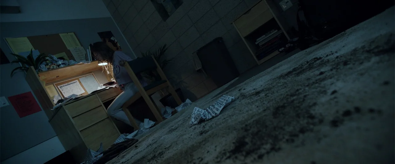 Nocturne (2020) movie still: wide — A dimly lit room features a student sitting at a cluttered wooden desk. Papers are scattered on the carpeted…