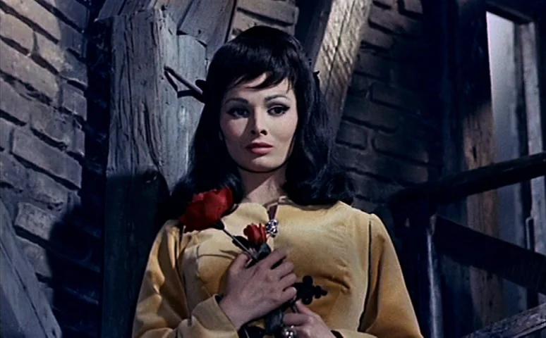 Mill of the Stone Women (1960) movie still: medium — A dimly lit indoor stairwell features dark brick walls and heavy wooden beams. A young woman in…