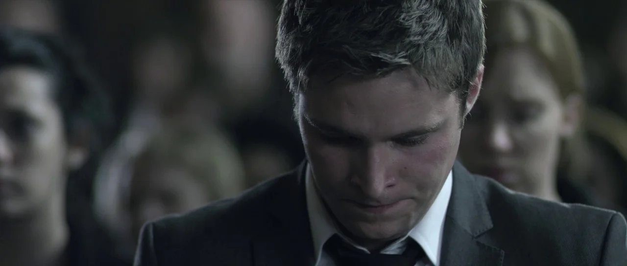 What Richard Did (2012) movie still: closeup — A man in a suit stands among a large, out-of-focus crowd of people. The man looks downward while…
