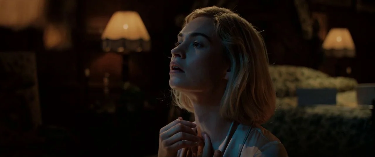 Rebecca movie still: closeup — A dimly lit room with vintage furniture and patterned upholstery in the background. A woman in a light robe looks up…