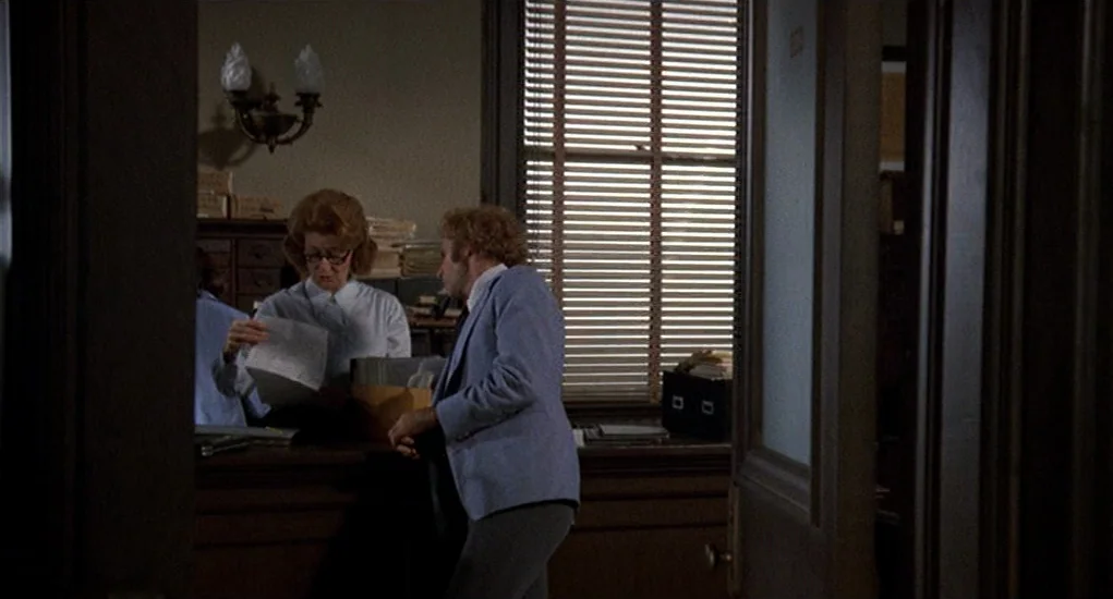 Family Plot (1976) movie still: medium — An administrative workspace features wooden cabinets and windows covered by horizontal venetian blinds. A…