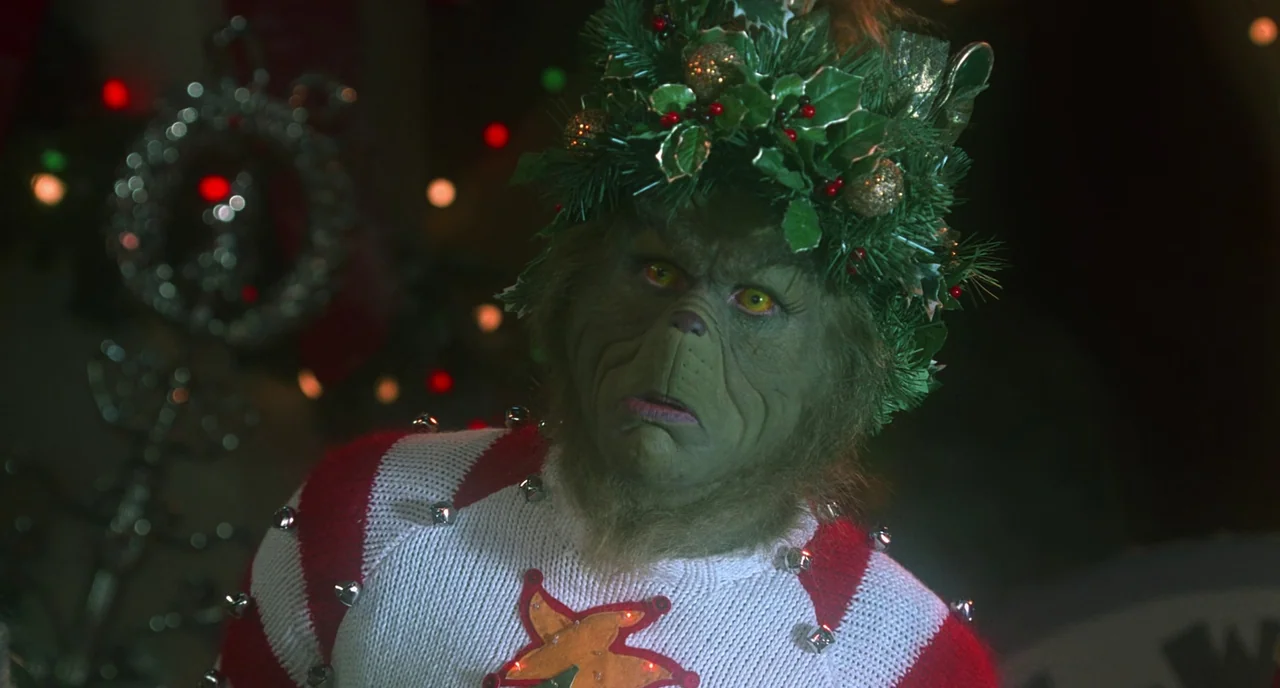 How The Grinch Stole Christmas (2000) movie still: medium — A green-skinned humanoid character with a holly wreath on his head stands indoors. He…