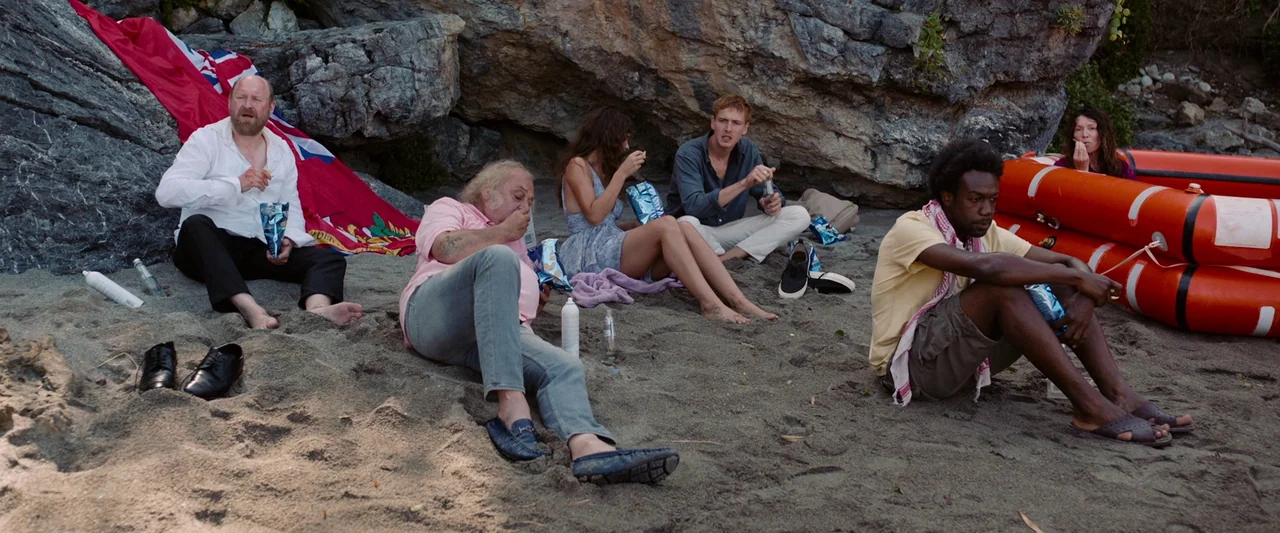 Triangle of Sadness (2022) movie still: wide — A group of six people sits on a sandy beach below a rocky ledge. They are resting near a red flag and…