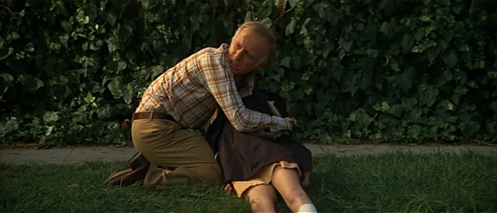 Assault on Precinct 13 (1976) movie still: medium — A man in a plaid shirt kneels on a grassy area next to a hedge. He clutches a second person who…