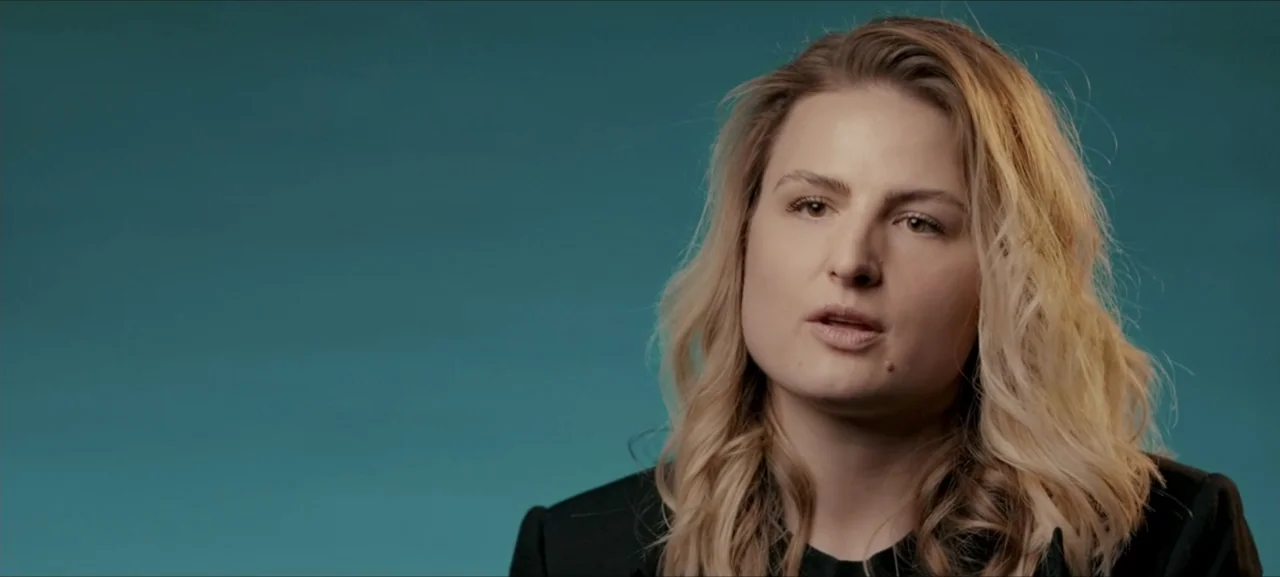 No Woman No Try (2022) movie still: medium — A studio environment features a plain solid teal backdrop behind the subject. A woman in a black blazer…