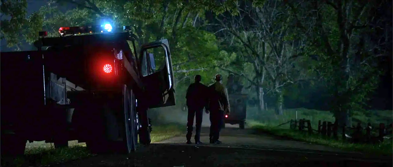 Jeepers Creepers 3 (2017) movie still: wide — A dark rural road is flanked by dense, dark trees at night. Two men walk away from the viewer towards…