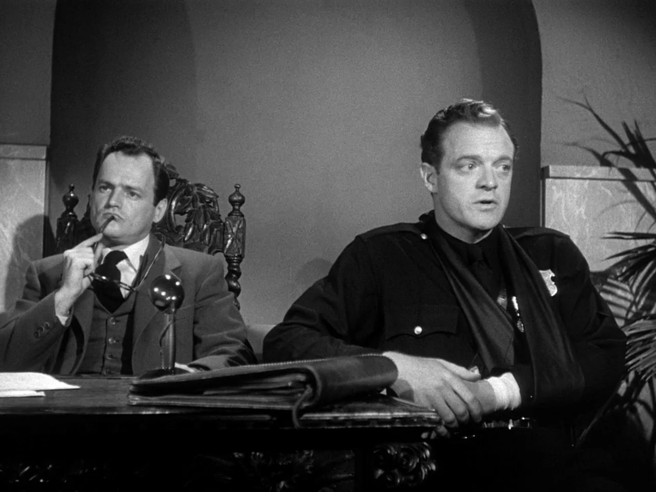 The Prowler (1951) movie still: medium — A formal office with a heavy wooden desk and an ornate carved wooden chair. A man in a suit sits beside a…