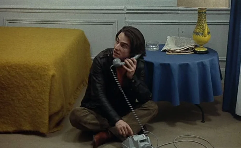 Bed & Board (1970) movie still: medium — A room contains a mustard yellow bed and a small table with a lamp. A man sits on the floor in front of the…