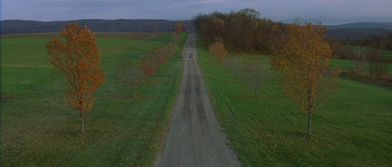 The Cider House Rules movie still: establishing — A long, straight gravel road stretches through vibrant green rolling hills and deciduous trees. A…