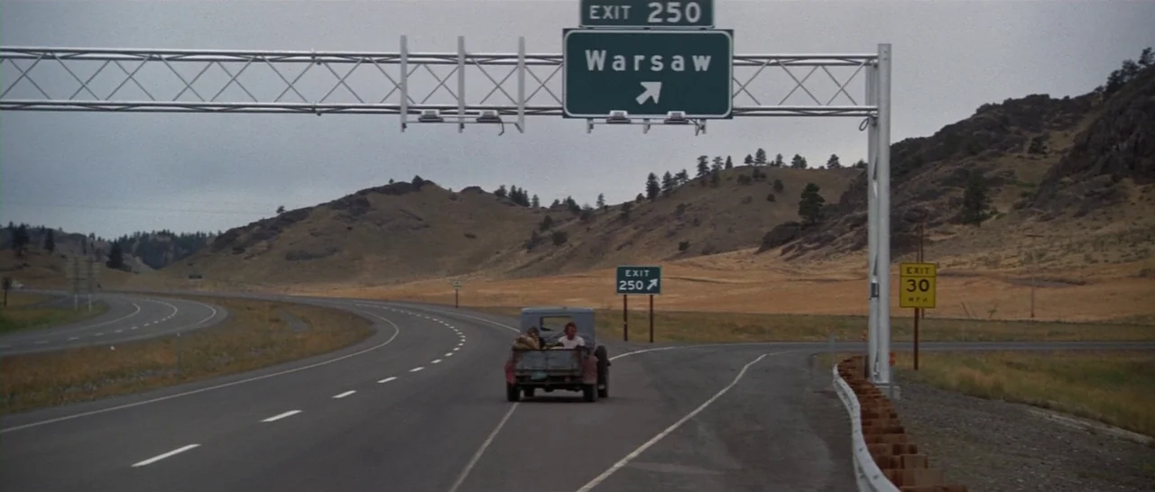Thunderbolt & Lightfoot (1974) movie still: wide — A rural highway stretches under a large metal overhead sign indicating an exit ramp. A vintage…
