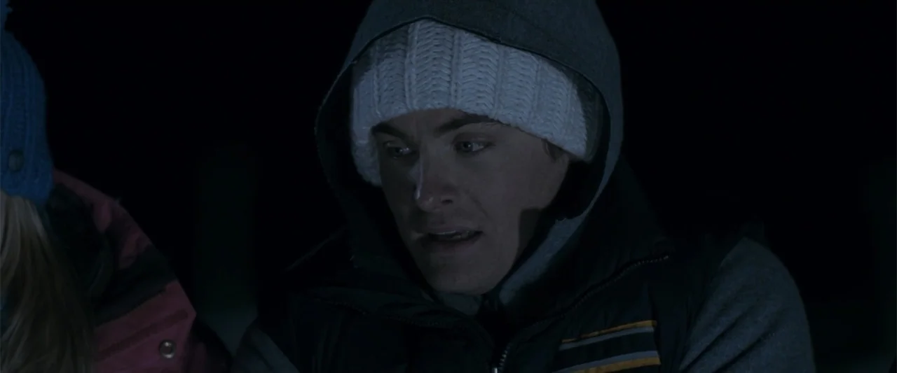 Frozen (2010) movie still: closeup — A dark outdoor setting at night features a man lit by a nearby fire. He wears a knit hat and hoodie while…