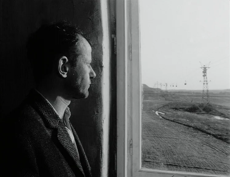 Damnation (1988) movie still: medium — A man stands inside a dimly lit room beside a tall rectangular window. He gazes out at a rural landscape…