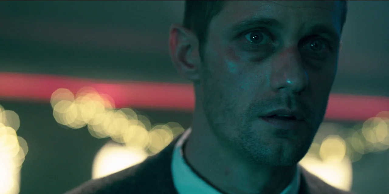 Mute (2018) movie still: closeup — A dark environment filled with out-of-focus golden lights and faint red neon lines. A man in a suit looks toward…