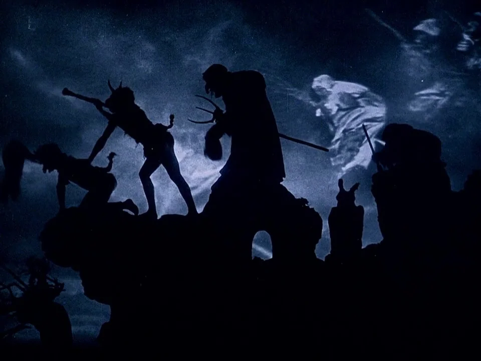Häxan (1922) movie still: wide — A dark, craggy rock formation silhouetted against a deep blue, cloudy night sky. Several figures in demonic attire…