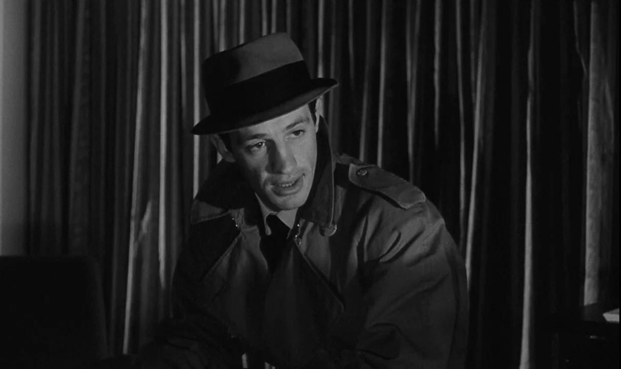 Le Doulos (1962) movie still: A striking medium-closeup in black and white captures a man in a fedora and trench coat, illuminated by a harsh side…