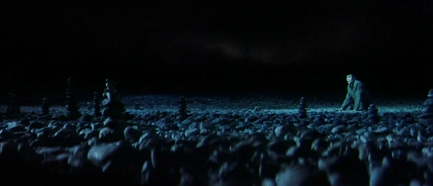 Jigoku (1960) movie still: wide — A dark, expansive rocky beach is covered with numerous small stone towers in the foreground. A lone man in a dark…